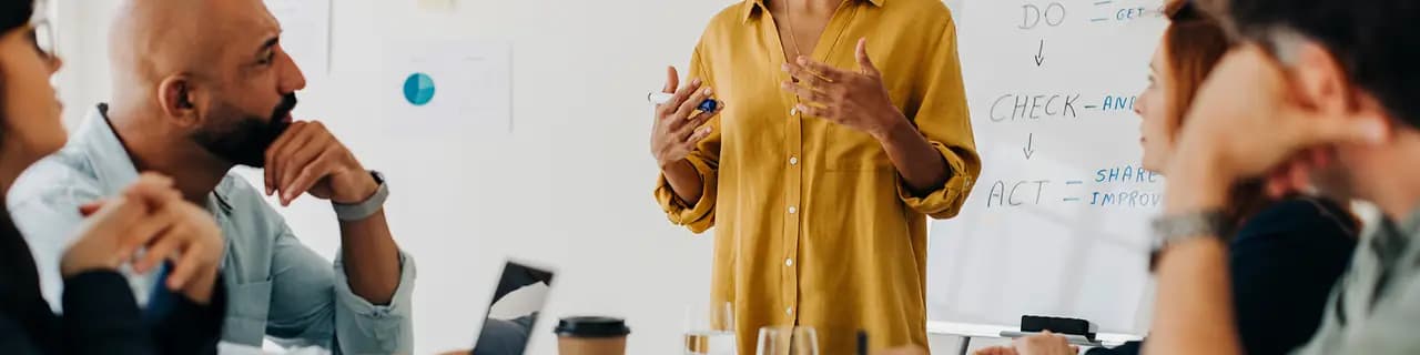 Une femme présente devant un tableau blanc portant les mots « DO », « CHECK », « ACT » ; plusieurs personnes l'écoutent.